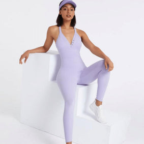 A woman models the Miranda Onesie and a matching lavender cap with white sneakers, striking a pose with one leg up on a white geometric prop. Her stylish look is ideal for everyday wear against a simple white background.