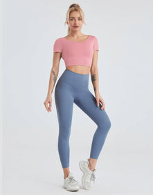 A woman stands against a plain light background in a chic outfit: SoHo Leggings with a high waist and medium-weight fabric, a pink cropped top, and white sneakers. Her blonde hair is tied back and she has tattoos on both arms.